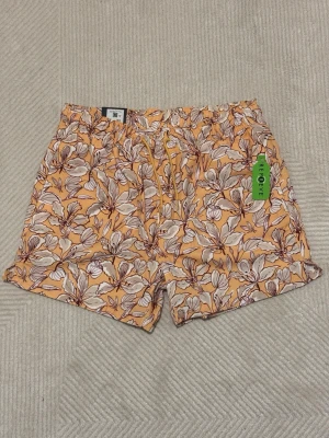 Blommiga badshorts från Dressmann - Säljer ett par snygga badshorts från Dressmann i storlek L. De har ett blommigt mönster i beige, brun och gul med dragsko i midjan och meshfoder. Perfekta för strand eller poolhäng!