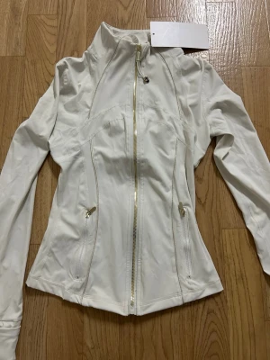 Vit vindjacka från Lululemon - Snygg vit vindjacka från Lululemon med figurnära passform och guldfärgade dragkedjor. Jackan har hög krage, långa ärmar och två fickor med dragkedja framtill. Materialet är mjukt och stretchigt, perfekt för en aktiv vardag.