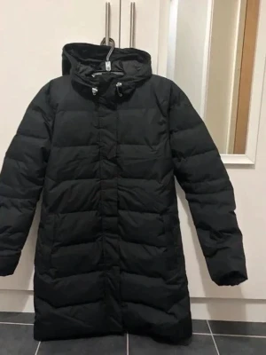 Svart lång pufferjacka Helly Hansen - Säljer en svart lång pufferjacka från Helly Hansen med huva och fuskpälsdetalj. Jackan har dragkedja framtill, snörning i huvan och stora fickor. Perfekt för kalla dagar och riktigt skön att bära. Klassisk och stilren design.