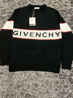 Svart stickad tröja från Givenchy - Säljer en svart stickad tröja från Givenchy med vit panel och röd kant över bröstet där loggan är tydligt broderad. Tröjan har långa ärmar och ribbade muddar vid hals, ärmslut och nederkant. Perfekt för dig som gillar exklusiva streetwear-plagg.