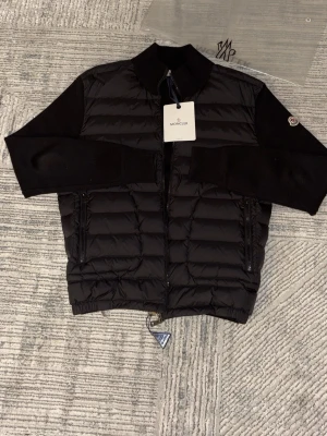 Svart dunväst från Moncler - Säljer en svart dunväst från Moncler med quiltad framsida och stickade ärmar. Västen har dragkedja framtill, två sidofickor med dragkedja och en hög krage. Klassisk Moncler-logga på ärmen och stilren design som passar till många looks.