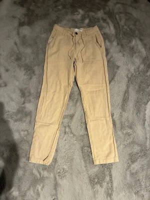 Beiga linnebyxor från Selected Homme - Snygga beiga linnebyxor från Selected Homme med avslappnad passform och dragsko i midjan. Byxorna har både fram- och bakfickor samt knappstängning. Perfekta för varma dagar och ger en chill vibe till din outfit.