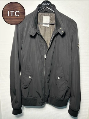 Fieldjacket från J.Lindeberg - Snygg svart fieldjacket från J.Lindeberg med klassisk krage, dragkedja framtill och två fickor med knappstängning. Jackan har ribbade muddar vid ärmslut och midja samt en diskret logga på ärmen. Perfekt för dig som vill ha en stilren och enkel look.