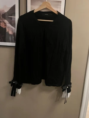 Svart kofta med rosetter och pärldetalj - Säljer en svart kofta från Vero Moda med långa ärmar. Ärmsluten har vita plisserade detaljer, svarta rosetter och pärldekorationer 