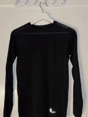 Svart långärmad träningströja Ronhill - Säljer en svart långärmad träningströja från Ronhill. Tröjan har en enkel och sportig design med rund hals och diskret logga nere vid fållen. Perfekt för löpning eller gympass. Tillverkad i lätt och snabbtorkande syntetmaterial.