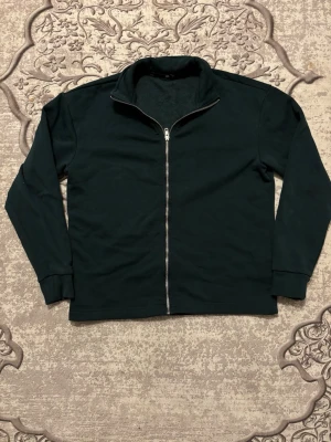 Mörkgrön/oliv zip hoodie från 157, storlek S - En mörkgrön/oliv hoodie från 157 i storlek S med hel dragkedja framtill och hög krage. Tillverkad i mjuk bomull med fleecefoder på insidan för extra komfort. Perfekt för kyliga dagar och enkel att matcha med allt.