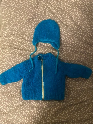 Blå stickad väst med dragkedja - Säljer en blå stickad väst med grov struktur och dragkedja framtill. Västen har en rund ring som dragkedjehänge och matchande blå mössa med knytband. 