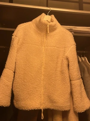 Beige teddyjacka med dragkedja - Mysig beige teddyjacka med hög krage och hel dragkedja framtill. Jackan har en fluffig och mjuk struktur som ger en chill vibe. Perfekt för kalla dagar när du vill vara både varm och snygg. Passar till det mesta och är lätt att matcha.