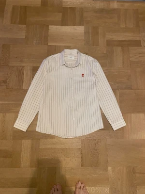 Randig skjorta från Ami Alexandre Mattiussi - Large - Säljer en vit skjorta från Ami Alexandre Mattiussi med tunna svarta ränder och röd broderad logga med hjärta på bröstet. Klassisk krage, långärmad och knäppning framtill. Skjortan är i bomull och har en clean, stilren vibe.
