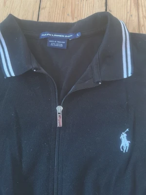 Svart pikétröja från Ralph Lauren Golf - Klassisk svart pikétröja från Ralph Lauren Golf med dragkedja framtill och vit broderad logga på bröstet. Kragen har vita ränder och tröjan är gjord i bomull med lite elastan för extra komfort. Perfekt till en sportig och stilren look.