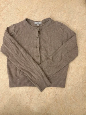 Beige kofta från H&M, storlek S - Mysig beige kofta från H&M i storlek S. Klassisk modell med rund halsringning, ribbade muddar och knappar framtill. Perfekt att slänga över en t-shirt eller linne för extra lager. Enkel och stilren design som funkar till det mesta.