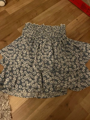 Blommig blå kortkjol med volanger - Supersöt blå kortkjol med vita blommor och volangkant. Kjolen har smock i midjan för en bekväm passform och ett luftigt, flowy tyg som passar perfekt till varma dagar. Perfekt att styla med en enkel topp och sneakers.