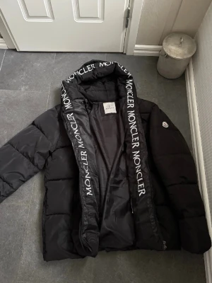 Svart Moncler pufferjacka med huva - Säljer en svart pufferjacka från Moncler med stor huva och logotyptext längs kanten. Jackan har dragkedja och är vadderad för extra värme. Perfekt för kalla dagar och har en clean, oversized look. Klassisk Moncler badge på ärmen. OBS endast använd en gång så näst intill nyskick! 