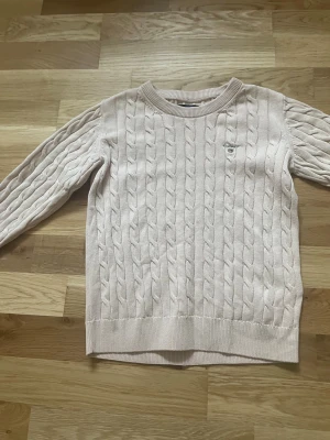 Rosa/begie  kabelstickad tröja från Gant - Kabelstickad Rosa/beige  tröja från Gant med rund halsringning och diskret logga på bröstet. Tröjan har långa ärmar och ribbade muddar vid ärmslut och nederkant. Perfekt för lager på lager under höst och vinter.