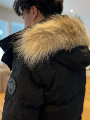 Svart pufferjacka från Canada Goose - Säljer en svart pufferjacka från Canada Goose med stor huva och avtagbar pälsdetalj. Jackan har diskret logga på ärmen, dragkedja och knappar framtill samt två sidofickor. Perfekt för kalla dagar och riktigt snygg till vinterlooken.