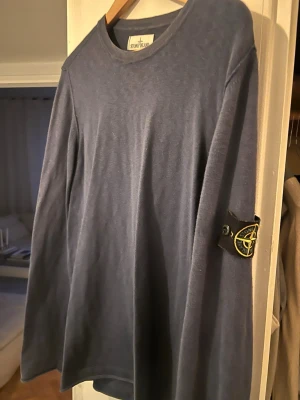 Blå långärmad tröja från Stone Island - Säljer en stilren blå långärmad tröja från Stone Island med den klassiska patchen på ärmen. Tröjan har rund halsringning och är gjord i mjuk bomull, perfekt för dig som gillar clean och enkel streetwear.