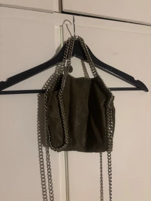 Grön axelväska Stella McCartney - Säljer en snygg grön axelväska från Stella McCartney med kedjeremmar och detaljer i silver. Väskan är gjord i mjuk mocka och har ett innerfack samt magnetknäppning. Perfekt storlek för det viktigaste och ger en cool touch till din outfit.