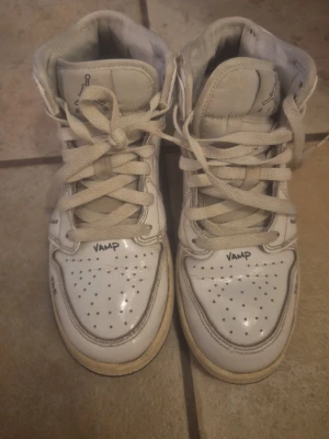 Nike Air Jordan 1 Mid vit/svart - Säljer ett par Nike Air Jordan 1 Mid i vitt med svarta detaljer och handskrivna markeringar på olika delar av skon. Klassisk hög siluett med snörning, perforerad tåbox och svart sula. Tillverkade i skinn och textil, perfekta för dig som gillar streetwear. Skorna har två små hål på sulan som man ser i bilderna.