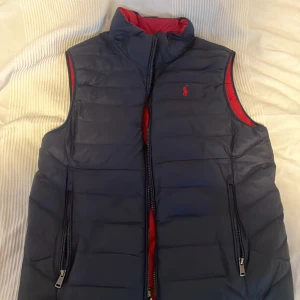 Blå dunväst från Polo Ralph Lauren - Snygg blå dunväst i skick 10/10 från Polo Ralph Lauren med röd insida och dragkedja framtill. Västen har två praktiska fickor med dragkedja och en liten logotyp på bröstet. Perfekt för kyliga dagar! Ny pris 2100