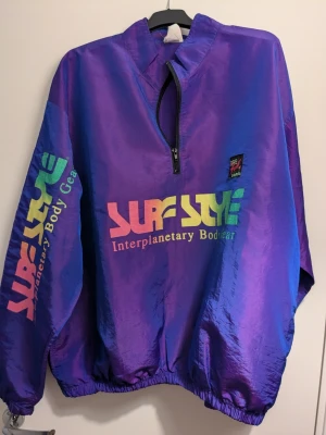 Lila Surf Style vindjacka med regnbågstryck - Retro vindjacka från Surf Style i glansig lila polyester med regnbågsfärgat tryck på bröst och ärm. Jackan har halv dragkedja, elastiska muddar och loose passform. Perfekt statement-plagg för dig som gillar färg och 90-talsvibbar.