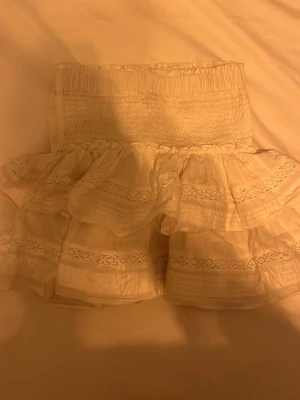 Vit volangkjol med spetsdetaljer - Supersöt kort vit kjol med bred smock i midjan och flera lager volanger. Kjolen har fina spetsdetaljer och ger en romantisk vibe. Perfekt för dig som gillar en feminin och luftig stil.