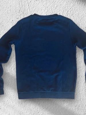 Mörkblå sweatshirt från Lacoste - Klassisk mörkblå sweatshirt från Lacoste med rund hals och det ikoniska krokodilmärket broderat på bröstet. Tröjan har långa ärmar och ribbade muddar vid ärmslut och nederkant. Perfekt för en clean och stilren look.