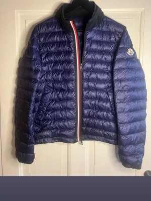 Blå Moncler dunjacka med inbyggd luva  - Säljer en mörkblå dunjacka från Moncler Longue Saison. Jackan har quiltad design, lång ärm och en snygg dragkedja med rödvit detalj. Klassisk Moncler-logga på ärmen och elastiska muddar. Perfekt för kyliga dagar. Kvitto finns då den köpt från merchsweden. Kom privat för frågor