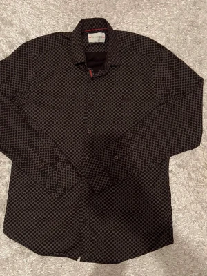 Svart mönstrad skjorta från Gucci - Snygg svart skjorta från Gucci med ikoniskt GG-mönster över hela tyget. Skjortan har klassisk krage, knäppning framtill och är tillverkad i 100% bomull. Perfekt för dig som vill ha en statement piece med lyxig känsla.