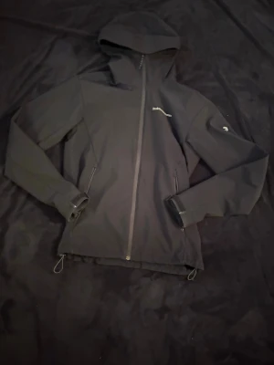 Svart vindjacka från Peak Performance - Snygg svart vindjacka från Peak Performance med huva och hel dragkedja framtill. Jackan har två fickor med dragkedja och justerbar nederkant. Perfekt för blåsiga dagar och enkel att matcha med olika outfits.