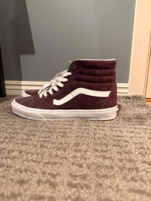 Vinröda Vans Sk8-Hi sneakers mocka - Säljer ett par Vans Sk8-Hi sneakers i vinröd mocka med klassisk vit sula och vit sidostripes. Skorna har vita snören och hög siluett som ger en retro vibe. Perfekta för dig som vill ha en clean och stilren look med lite edge.