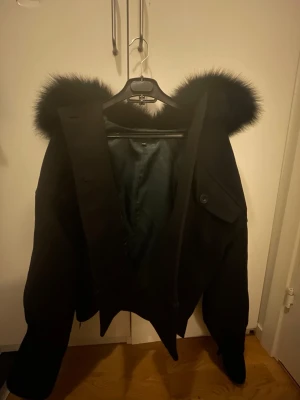 Svart kappa med fuskpälsluva - Säljer en svart kappa med stor luva kantad av fluffig fuskpäls. Jackan har en klassisk rak passform, knappar framtill och en ficka med lock på bröstet. Perfekt för dig som vill ha en stilren och varm look under kyliga dagar.