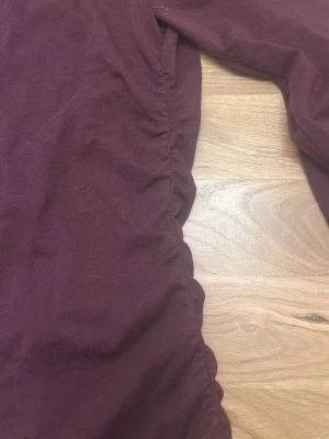 Vinröd offshoulder topp Gina Tricot - Säljer en vinröd offshoulder topp från Gina Tricot i mjukt bomullsmaterial. Toppen har långa ärmar och rynkade detaljer längs sidorna som ger en snygg form. Perfekt till jeans eller kjol för en trendig look.