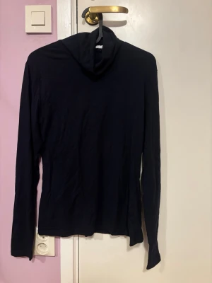 Svart polotröja  från Samsøe Samsøe - En enkel och stilren svart polotröja från Samsøe Samsøe i storlek M/L. Tröjan har långa ärmar, tillverkad i mjukt bomullsmaterial. Perfekt för dig som gillar minimalistisk och clean stil.