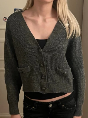 Mörkgrå stickad kofta med knappar - Säljer en mörkgrå stickad kofta med v-ringning och fyra stora knappar framtill. Koftan är croppad och har två dekorativa fickor med knappdetaljer. Perfekt till höst och vinter för en chill och snygg look.