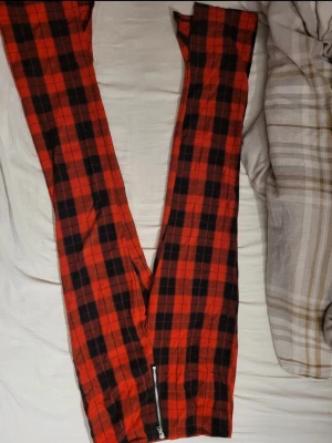 Rödrutiga byxor med dragkedja - Säljer ett par röda byxor med svart rutmönster och dragkedja vid benslutet. Byxorna har en smal passform och är perfekta för dig som vill sticka ut med en cool och trendig stil. Materialet känns mjukt och bekvämt, perfekt för vardagslooken.