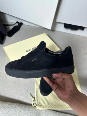 Svarta sneakers från Axel Arigato mocka - Säljer ett par svarta sneakers från Axel Arigato i mjuk mocka helt nya. Skorna har minimalistisk design med elastiskt band över vristen och diskret guldfärgad logga på sidan. Platt sula och rund tå ger en clean look. Perfekta till streetwear eller casual outfits. Kommer med box osv 