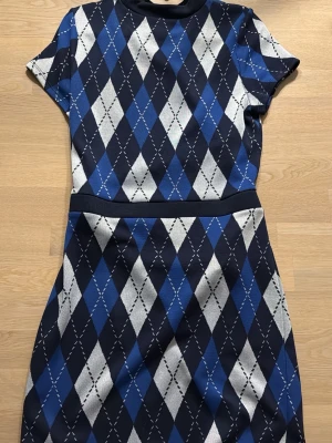 Blå och svart rutig kortklänning från Divided H&M i L  - Kortklänning med klassiskt argylemönster i blått, svart och vitt. Klänningen har rund hals, korta ärmar och markerad midja. Materialet känns mjukt och stretchigt, perfekt för en trendig och sportig look.