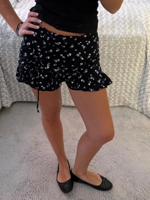 Blommig shortskjol - Supersöta svart shortskjol från Hollister med vitt blommönster. Modellen är högmidjad med volangkant och snörning i sidorna för extra detalj. Perfekta för sommaren och riktigt sköna att bära till crop tops eller linnen.