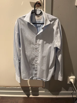 Ljusblå skjorta från The Shirt Factory - Klassisk ljusblå skjorta från The Shirt Factory i slim fit-modell. Skjortan är strykfri och har långa ärmar samt knappar framtill. Perfekt för dig som gillar stilrena och enkla plagg. Storleken är 38 som motsvarar typ S