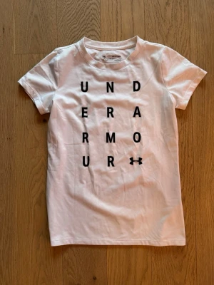 Vit Under Armour t-shirt XS-S - Snygg vit t-shirt från Under Armour i loose fit med svart tryck framtill där bokstäverna bildar märkesnamnet. Klassisk rund hals och korta ärmar. Tillverkad i mjuk bomull, perfekt för en sportig look.