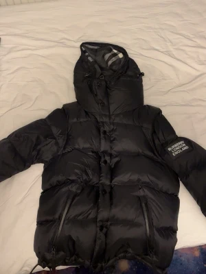Svart pufferjacka från Burberry - Säljer en svart pufferjacka från Burberry med ikoniskt rutigt foder i huvan. Jackan har dragkedjor på fickorna, knappar framtill och en patch med Burberry London England på ärmen. Perfekt för kalla dagar och riktigt snygg till streetwear.