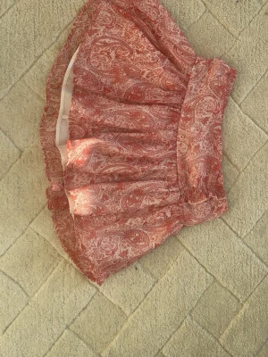 Rosa mönstrad kortkjol från Gina Tricot - Supersöt kortkjol från Gina Tricot i rosa och vitt med paisleyinspirerat mönster. Kjolen har resår i midjan och är gjord i ett lätt, luftigt material som känns perfekt för varma dagar. Passformen är normal och kjolen har en flowy look.