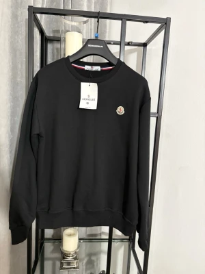 Svart sweatshirt från Moncler - Säljer en svart sweatshirt från Moncler med rund halsringning och Moncler-logga på bröstet. Tröjan har ribbade muddar vid ärmslut och nederkant samt en snygg detalj med röd, vit och blå rand på insidan av kragen. Perfekt för dig som gillar stilrena plagg.