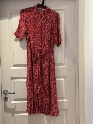 Röd blommig klänning från NA-KD - Röd midilång klänning från NA-KD med små blommor, knappar hela bägen och knytskärp i midjan. Klänningen har korta ärmar och klassisk krage, perfekt för vår och sommar. Klänningen har även fickor. Storlek 38 men oversize. 
