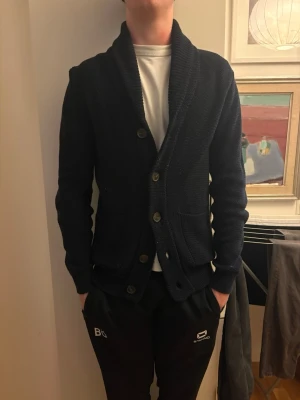 Stickad kofta Selected Homme - Mörkblå stickad kofta från Selected Homme med grovstickad struktur och bred sjalkrage. Koftan har stora knappar framtill och två praktiska fickor. Koftan är egentligen lite större än S, den funkar nog som en M också.Modellen är 180 cm.