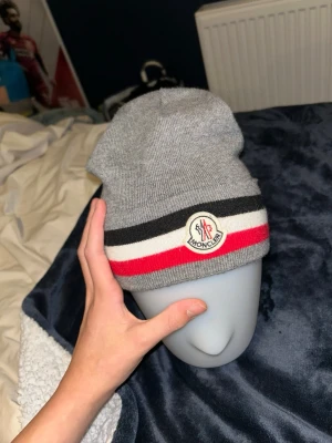 Grå Moncler mössa med ränder - Säljer en snygg grå mössa från Moncler med klassiska svarta, vita och röda ränder nertill. Mössan har Moncler-logga framtill och är stickad i mjukt material som passar perfekt för kyliga dagar.kom med bud vid intresse