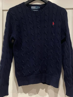 Mörkblå kabelstickad tröja Polo Ralph Lauren - Klassisk mörkblå kabelstickad tröja från Polo Ralph Lauren i bomull. Tröjan har rund hals, ribbade muddar och den ikoniska röda logon broderad på bröstet. Perfekt till lager på lager och funkar året runt.