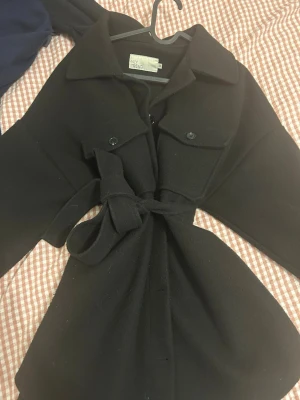 Svart kappa från NLY Trend med bälte - Snygg svart kappa från NLY Trend med klassisk krage, knappar framtill och två bröstfickor. Jackan har ett matchande bälte i midjan för en snygg siluett och är tillverkad i ett mjukt ullmix-material. Perfekt för kyligare dagar.