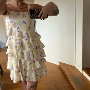 Blommig volangklänning - Säljer en superfin blommig klänning med volanger. Den är ärmlös med smala axelband och har en kort längd, perfekt för sommaren. Klänningen har ett härligt mönster i ljusa färger och är i nyskick. Perfekt för en somrig dag eller fest!