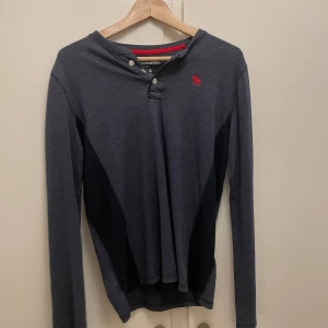 Mörkgrå långärmad tröja från Abercrombie Kids - Säljer en snygg mörkgrå långärmad tröja från Abercrombie Kids. Den har en liten röd logga på bröstet och knappar vid halsen för en cool look. Perfekt för höst och vinter, och passar både till vardags och lite finare tillfällen. Superbekväm och stilren!